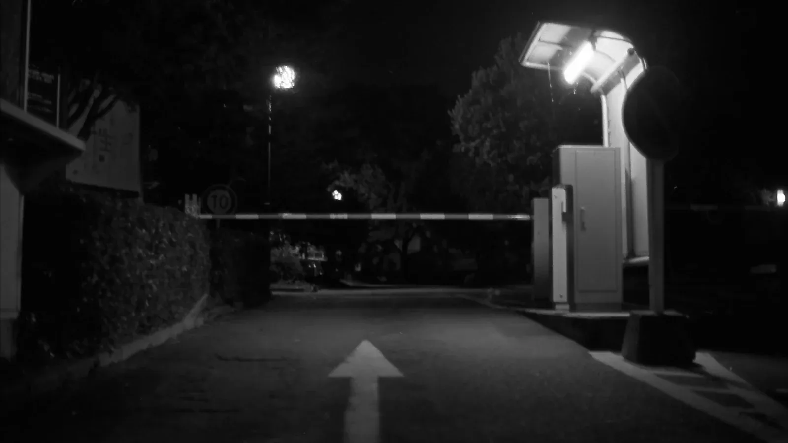 夜の構内ゲート。路面に手前を指す白い矢印、その奥に下りた縞模様の遮断バー、右に灯りの点いた警備ボックスと進入禁止標識、左は生垣と「10」の速度標識。2010年のモノクロフィルムスキャン