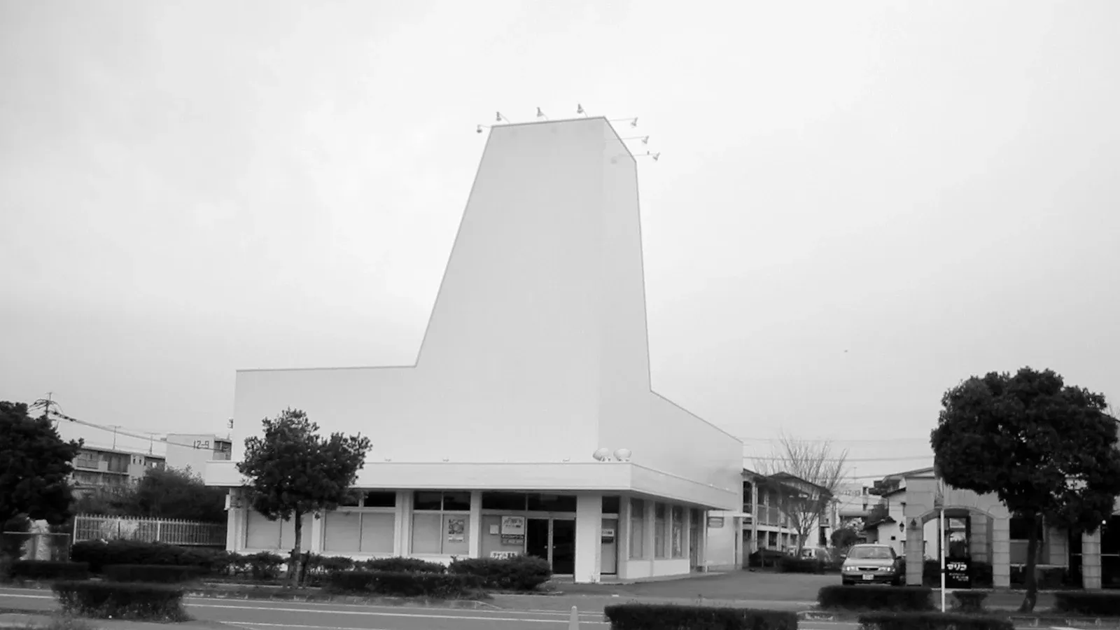 全国展開していた玩具店「ハローマック」の廃業後の姿。特徴的な城型の塔のファサードは看板を剥がされ、白く塗り潰されている。手前は空き地とカラーコーン、奥に住宅と曇り空。2002年のコンパクトデジカメで撮影